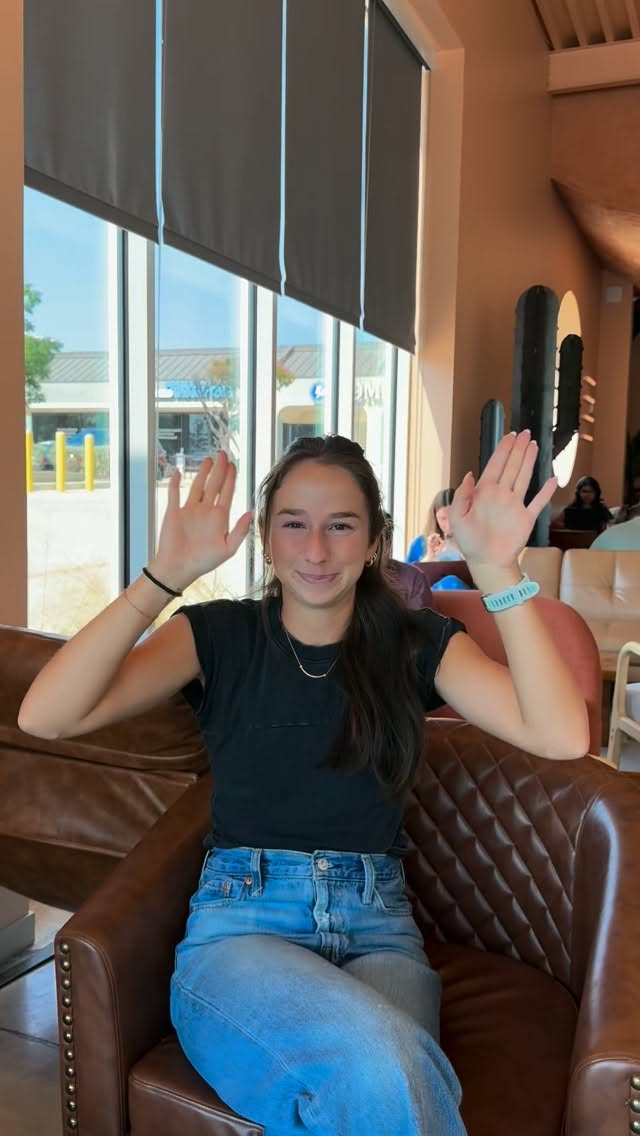 A young woman sits in a brown leather armchair in the Cliff Coffee lounge, smiling at the camera with both hands raised and palms facing forward. Tall windows behind her look out on the parking lot under a blue sky, and a dark cactus sculpture hangs on the terracotta wall.
