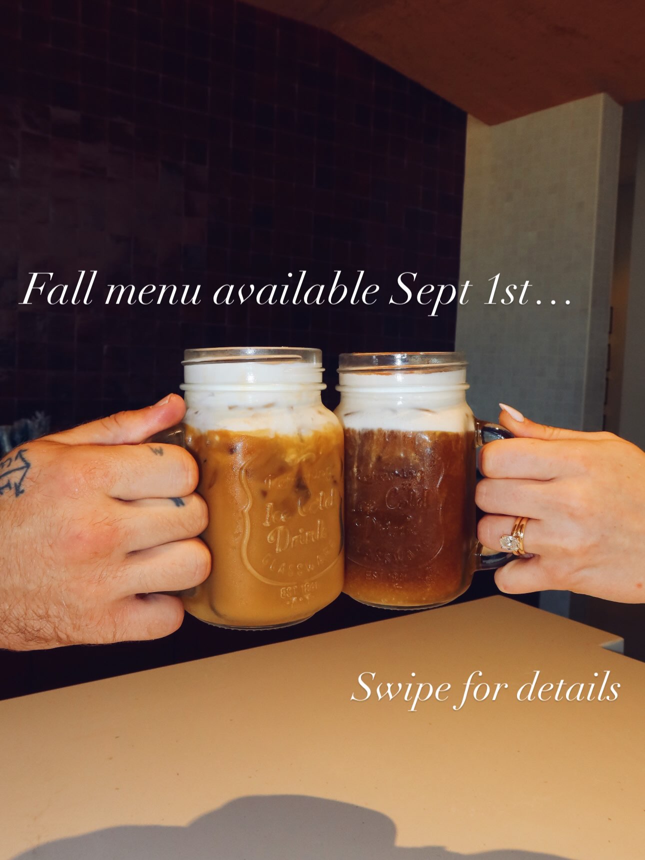 Two hands clink tall mason-jar mugs across the counter at Cliff Coffee. The left drink is a pale caramel-colored iced coffee under a thick layer of white foam; the right is much darker with cinnamon dusted over its foam. Text over the photo reads “Fall menu available Sept 1st…” and “Swipe for details.”