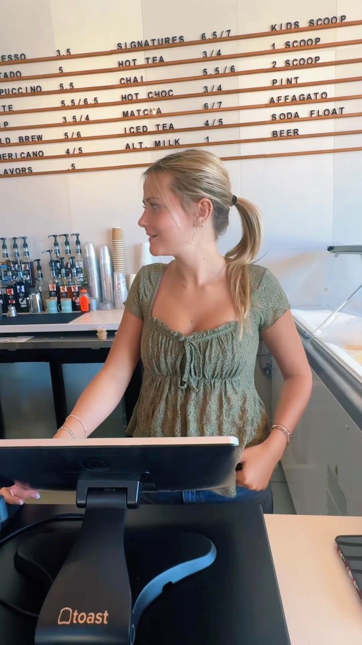A smiling barista behind the Cliff Coffee counter glances to one side at the Toast register, with the letter-board drink menu and the syrup station behind her.