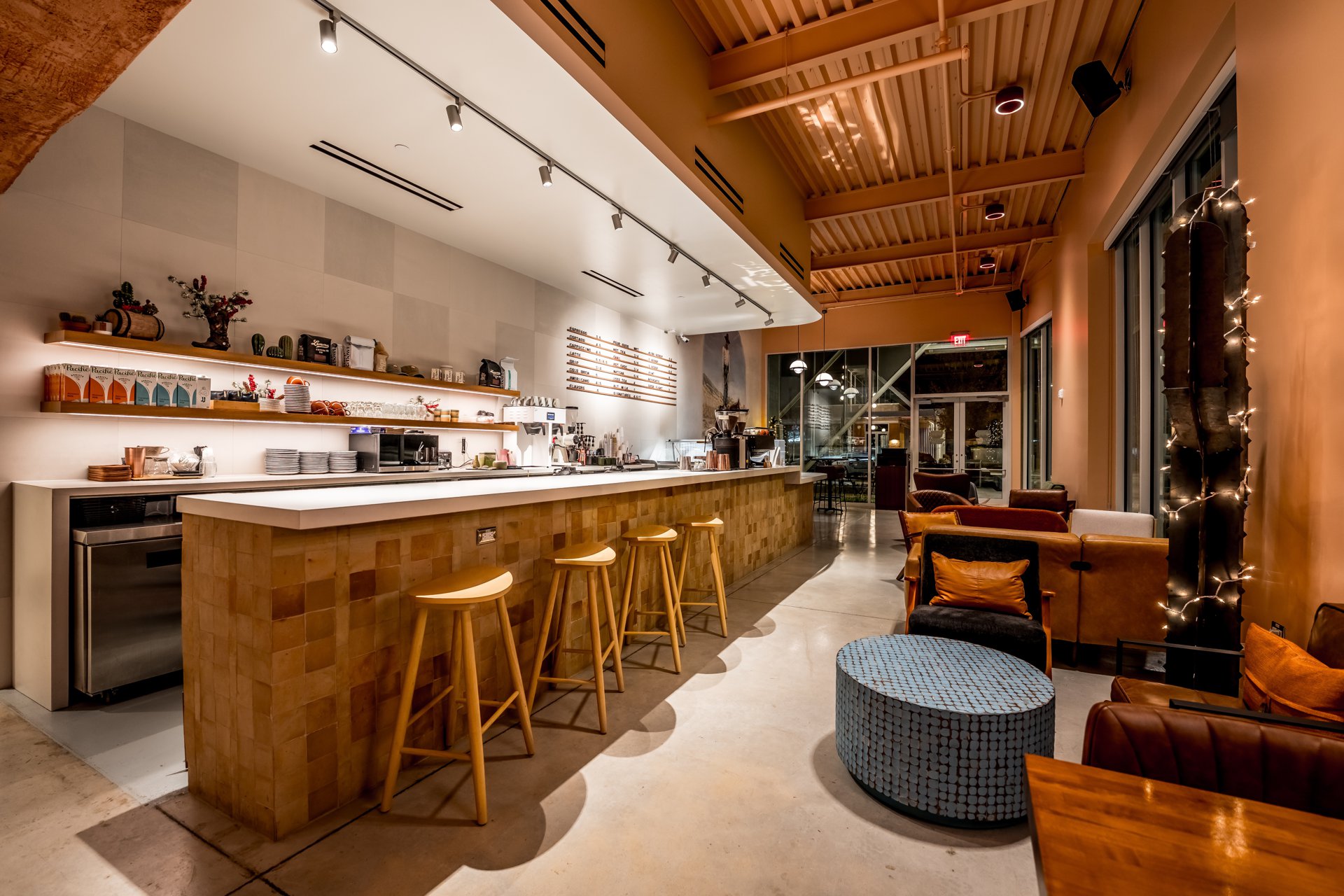 The bar inside Cliff Coffee: tile-fronted counter, wooden stools, shelves of retail and plants, with the lounge and string lights toward the windows