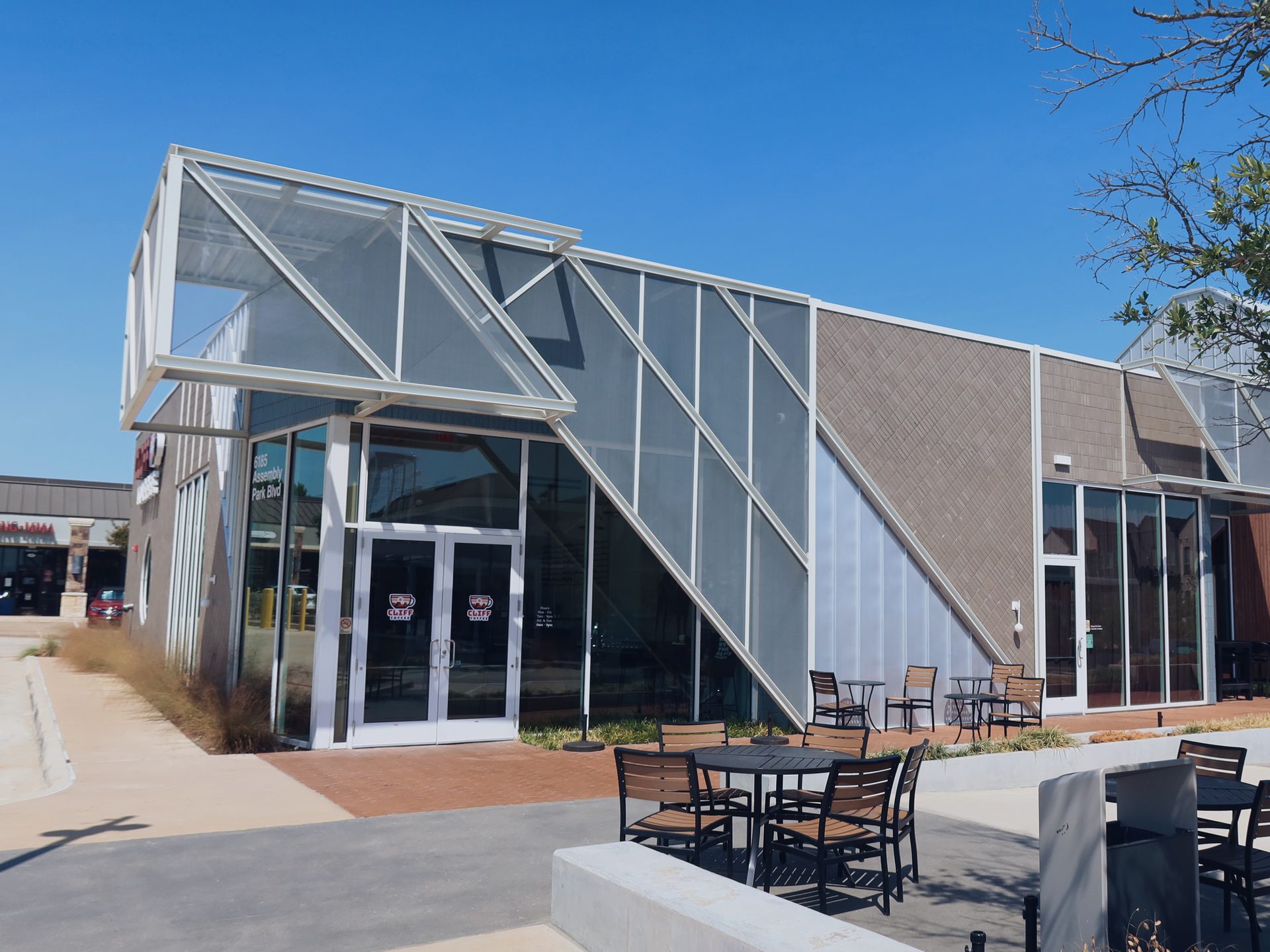 The Cliff Coffee storefront: glass double doors with the Cliff Coffee cup logo on each, under a white steel canopy, with patio tables to the right